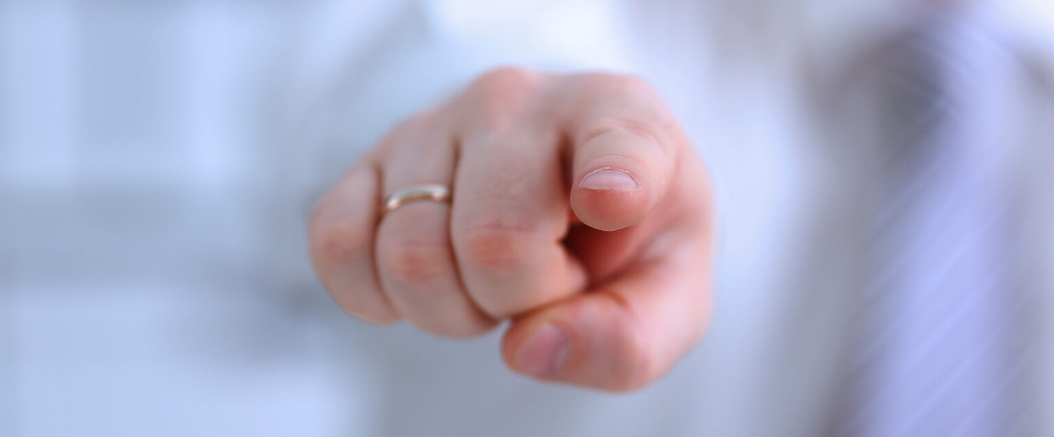 A male arm, clothed in white, reaches out to point his index finger at you. The white-skinned hand has a plain, gold wedding ring on its ring finger.