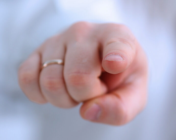 A male arm, clothed in white, reaches out to point his index finger at you. The white-skinned hand has a plain, gold wedding ring on its ring finger.