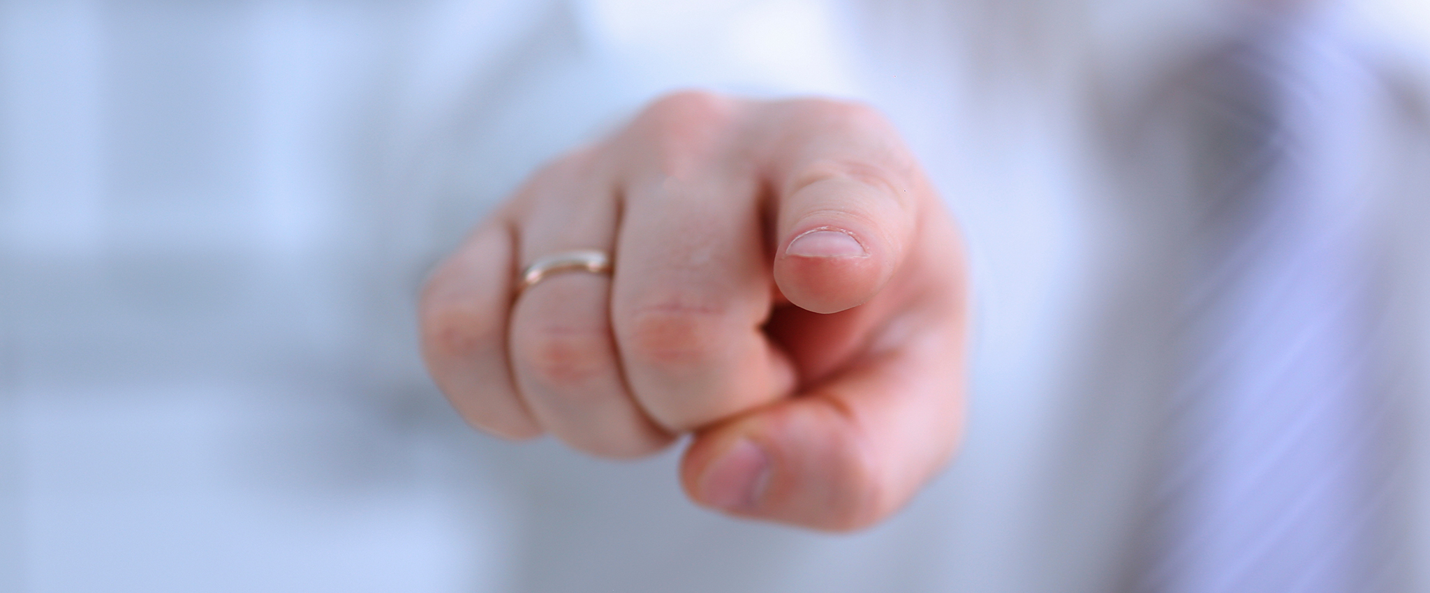 A male arm, clothed in white, reaches out to point his index finger at you. The white-skinned hand has a plain, gold wedding ring on its ring finger.