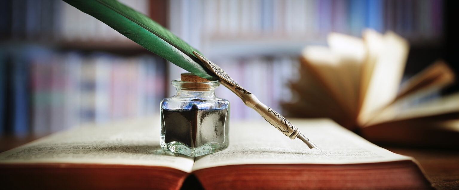 A quill pen and an ink well rest on an old, open book in a library.