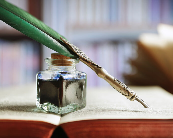 A quill pen and an ink well rest on an old, open book in a library.