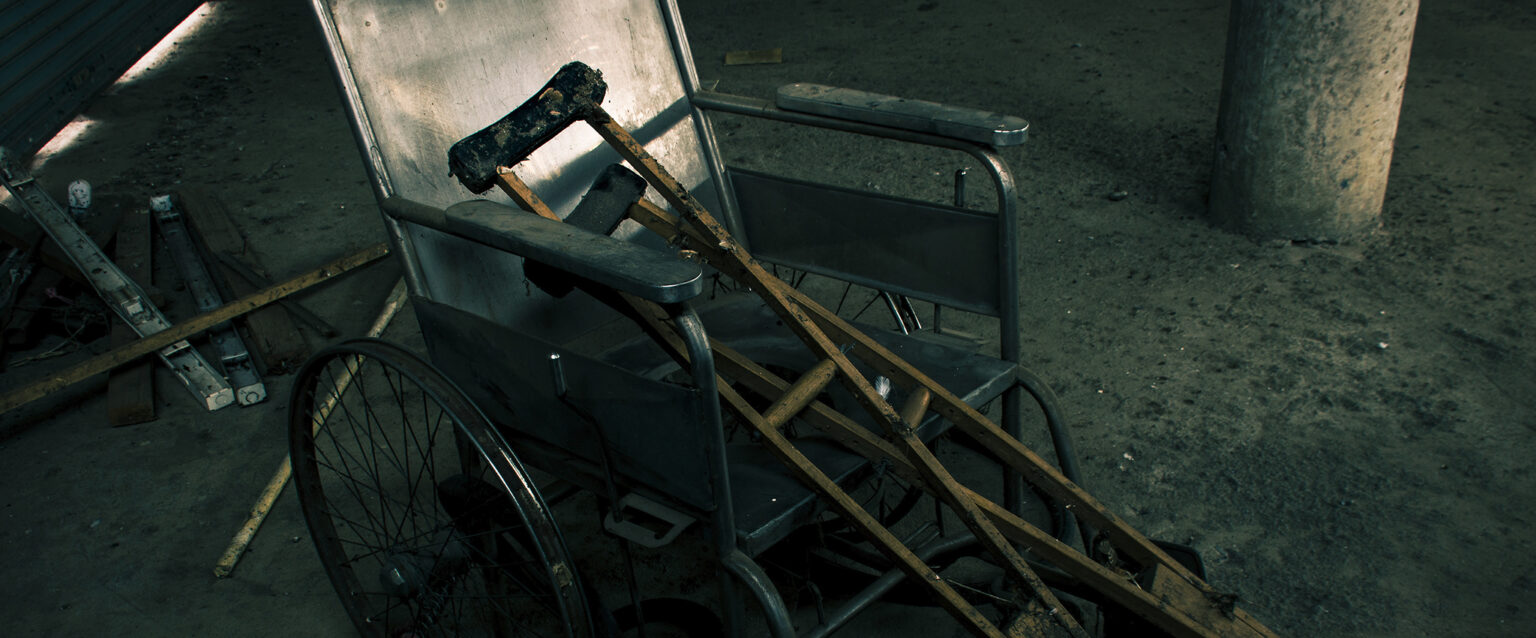 An old, abandoned wheelchair and wooden crutches on a basement cement floor with other abandoned items in a pile behind them.