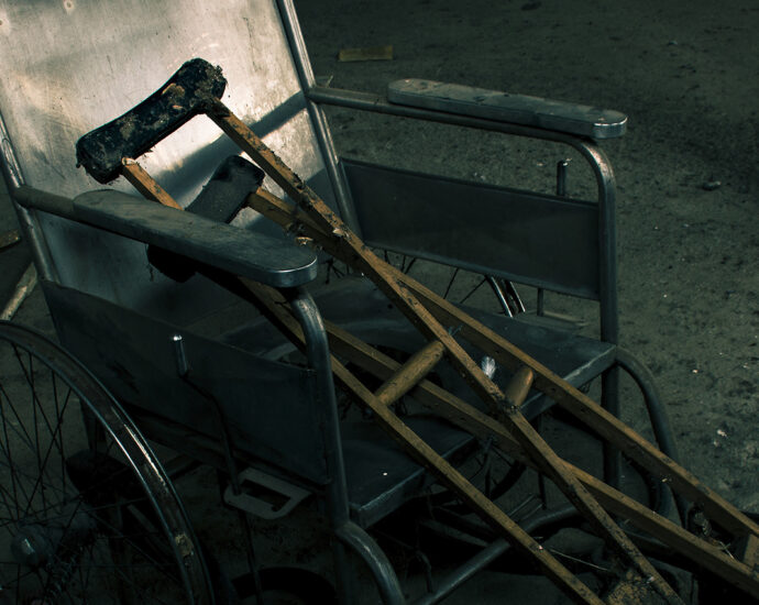 An old, abandoned wheelchair and wooden crutches on a basement cement floor with other abandoned items in a pile behind them.