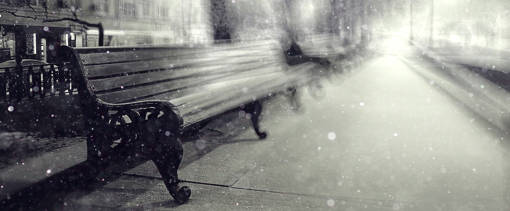 A monochrome image of snow falling softly on a line of wood and cast-iron benches that line the sidewalk in a New York City park. In the background, old buildings stretch into the distance.