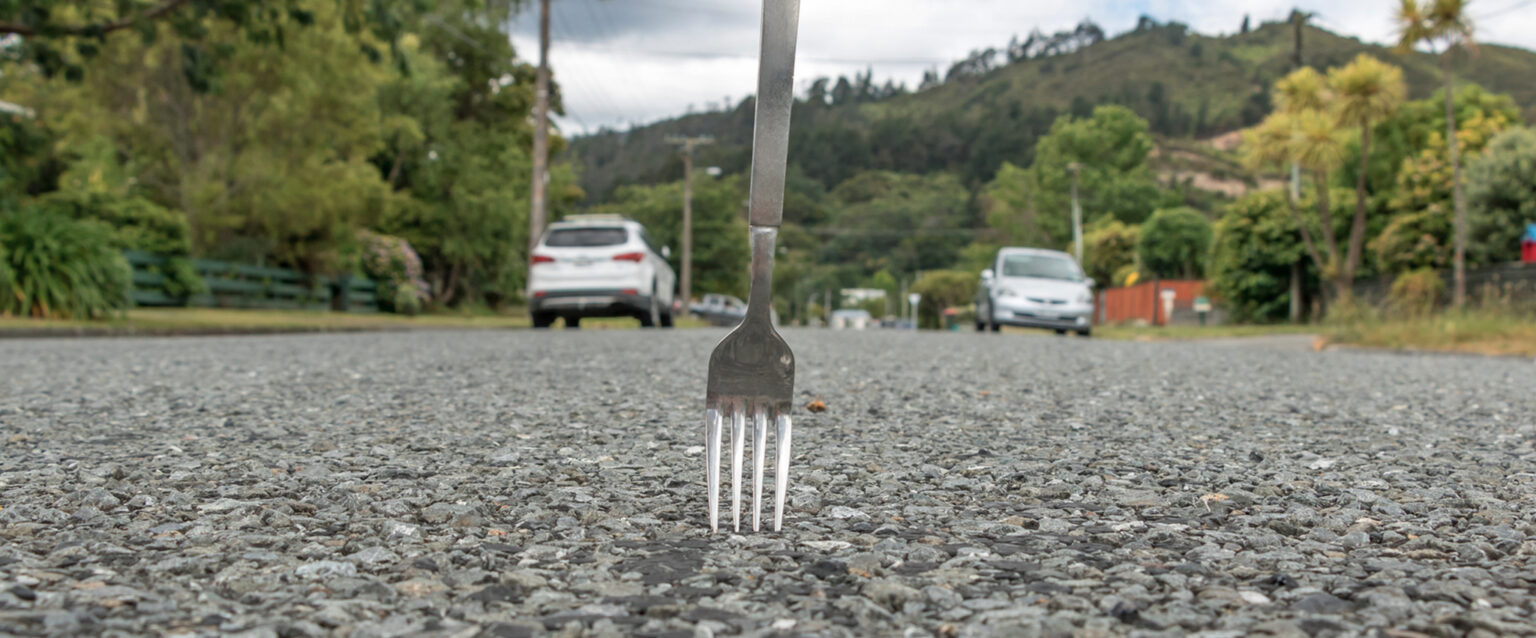 A fork stands atop a gravel road, near mountains. The tines are touching the tiny gravel beneath them. A few cars and a couple of fences can be seen in the distance, lining the road.