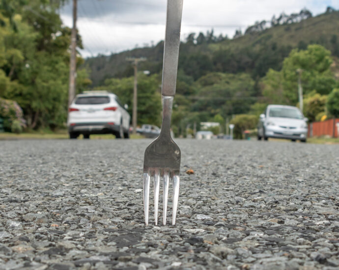 A fork stands atop a gravel road, near mountains. The tines are touching the tiny gravel beneath them. A few cars and a couple of fences can be seen in the distance, lining the road.