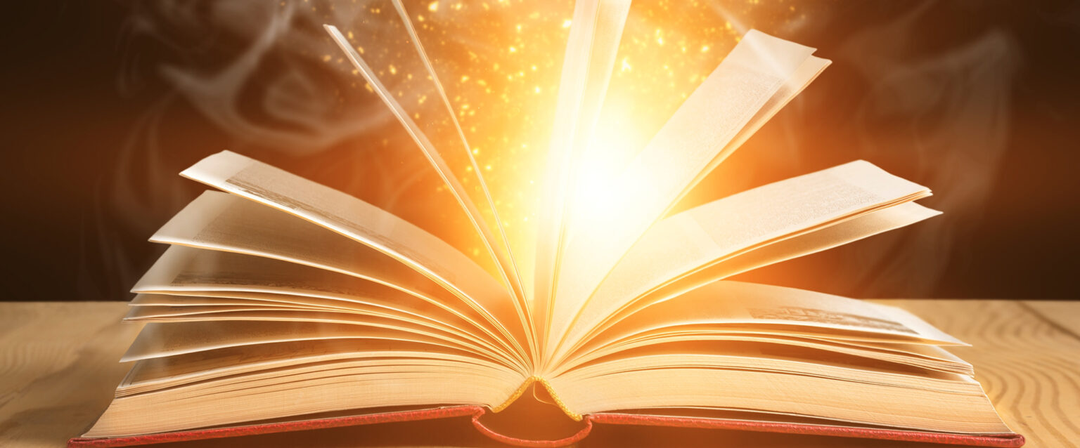 An open book, with pages extended upward, lies on a light-colored wooden desk. Sparkles, light, and smoke appear to be coming from within it.