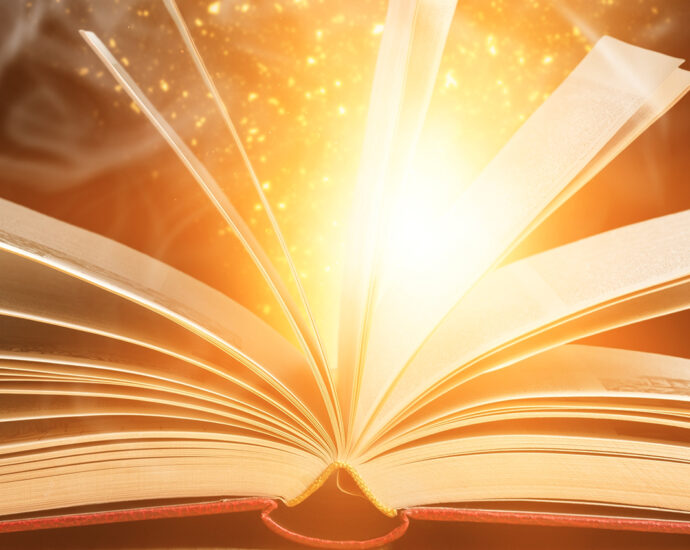 An open book, with pages extended upward, lies on a light-colored wooden desk. Sparkles, light, and smoke appear to be coming from within it.
