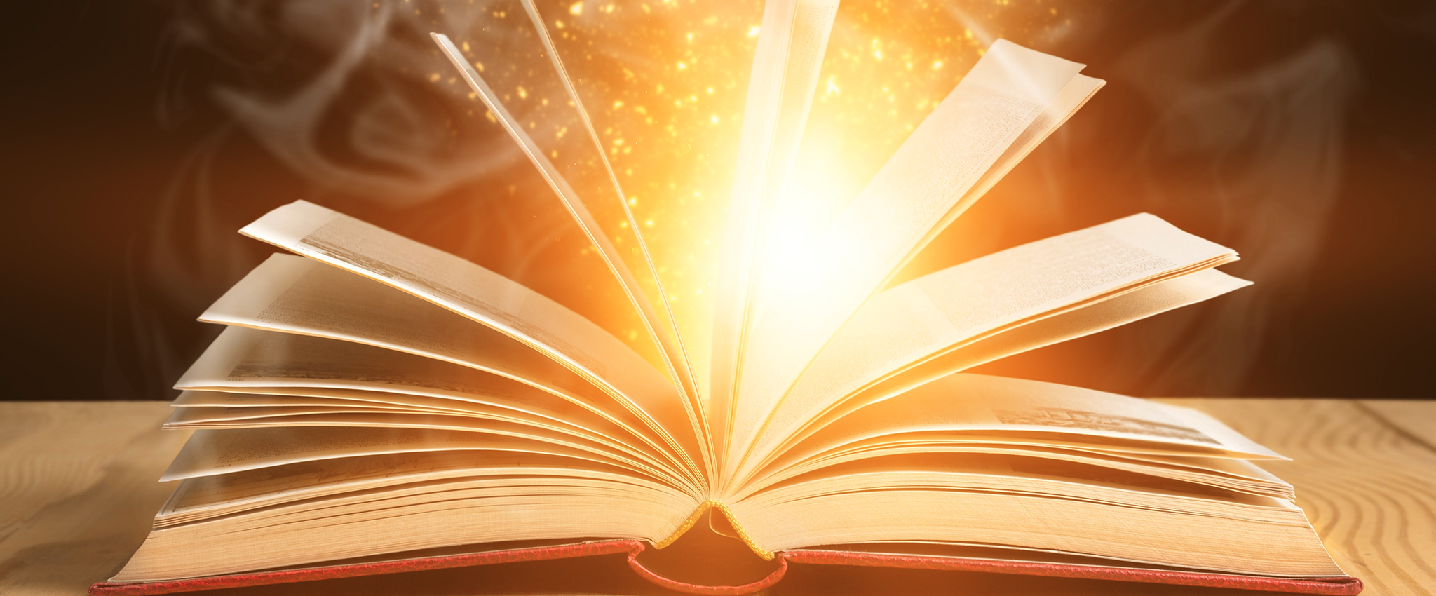 An open book, with pages extended upward, lies on a light-colored wooden desk. Sparkles, light, and smoke appear to be coming from within it.