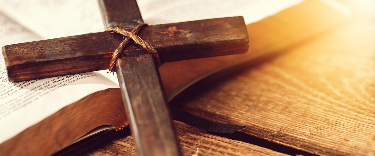 A wooden cross held together with crisscrossing twine lies across a wooden plank table and rests on an open Bible.