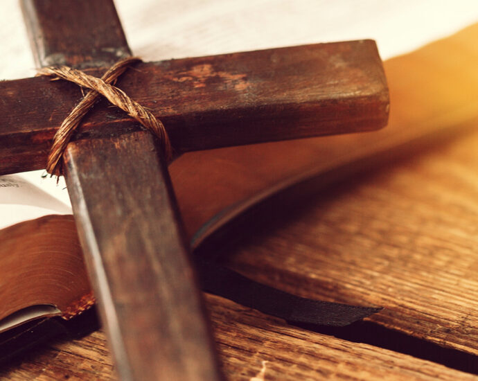 A wooden cross held together with crisscrossing twine lies across a wooden plank table and rests on an open Bible.