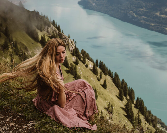 A woman in a light mauve dress sits on the side of a grass-covered mountain in Switzerland. At the base of the mountain is a clear, pale blue lake.