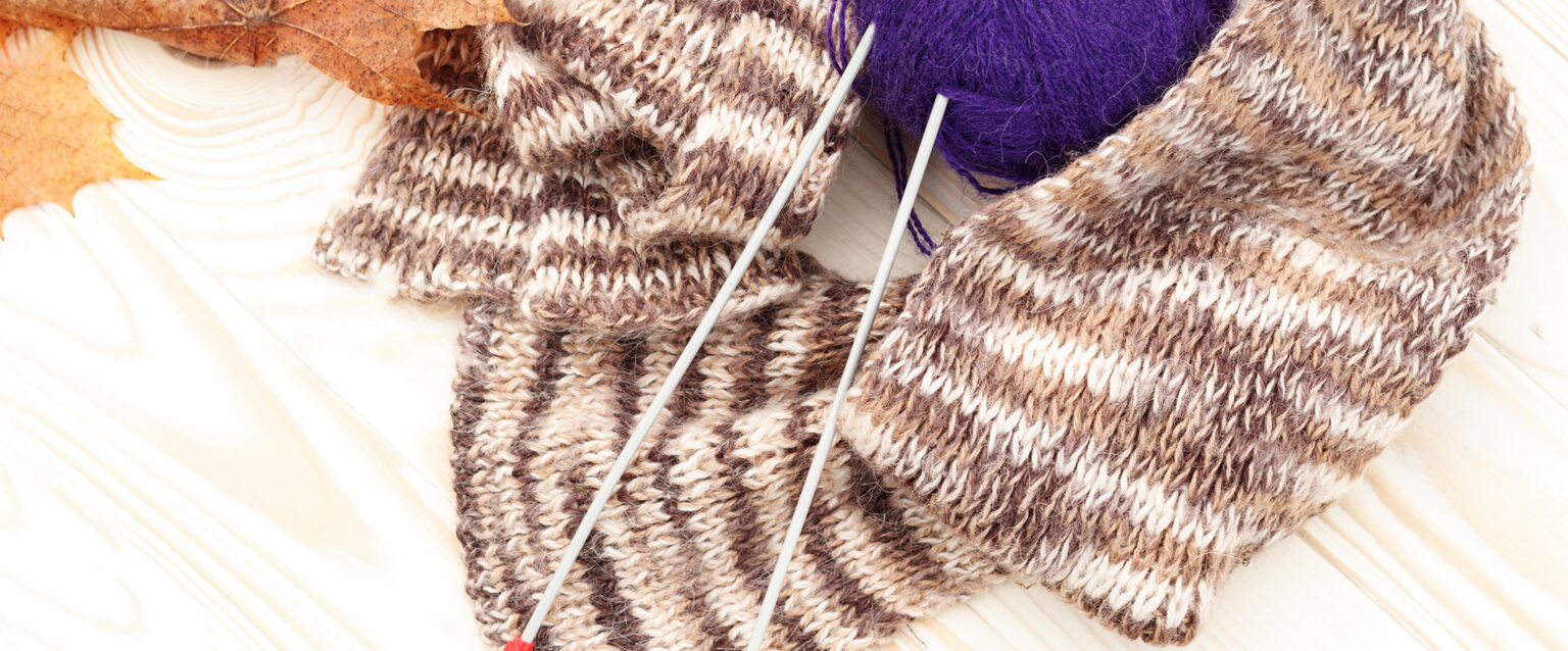 A purple ball of wool yarn, with two silver knitting needles poking through it, is nestled within a white, tan, and brown knit scarf, beside two browned leaves, on a white wooden table.