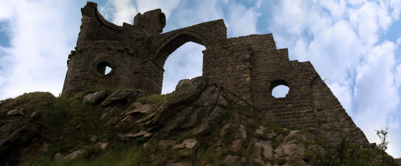 A single wall and turret of Mow Cop Castle as seen from below, against a cloudy blue sky, on a rocky hillside in the Biddulph valley in the county of Staffordshire.