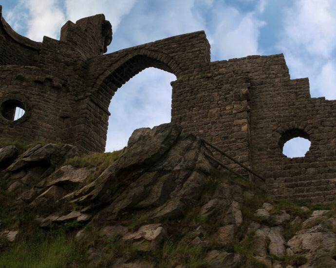 A single wall and turret of Mow Cop Castle as seen from below, against a cloudy blue sky, on a rocky hillside in the Biddulph valley in the county of Staffordshire.