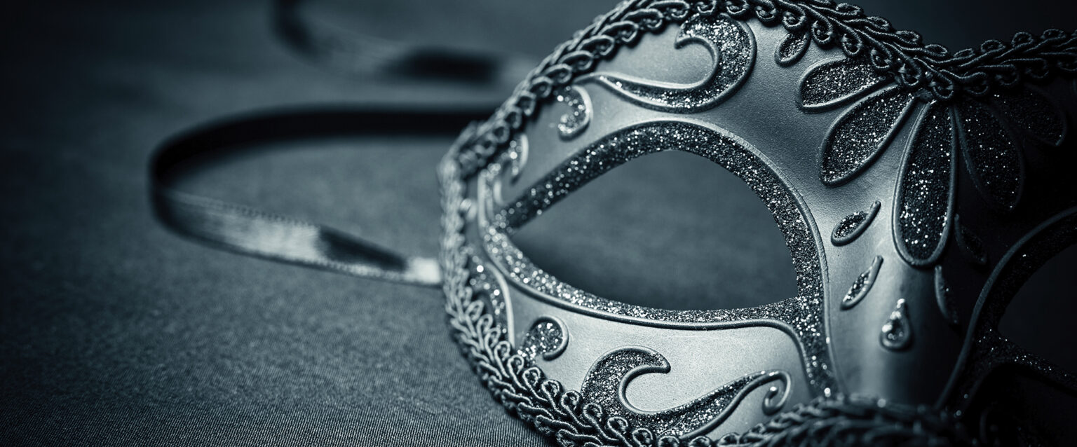 A monochrome image of a black carnival mask, with ribbon trailing, sits on material.