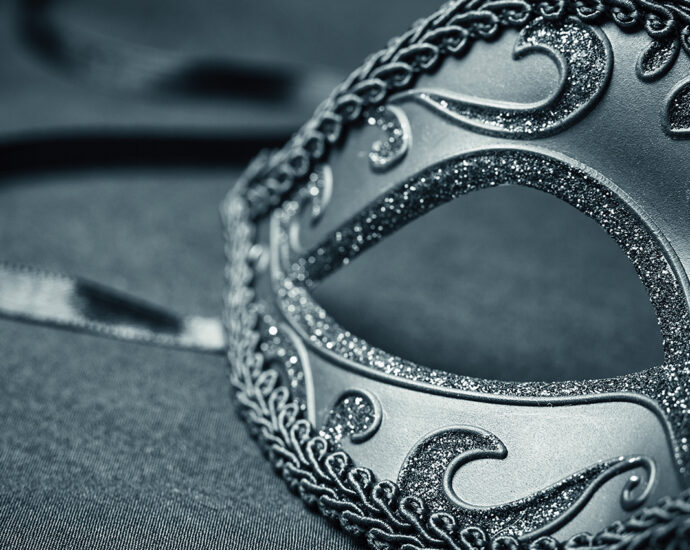 A monochrome image of a black carnival mask, with ribbon trailing, sits on material.