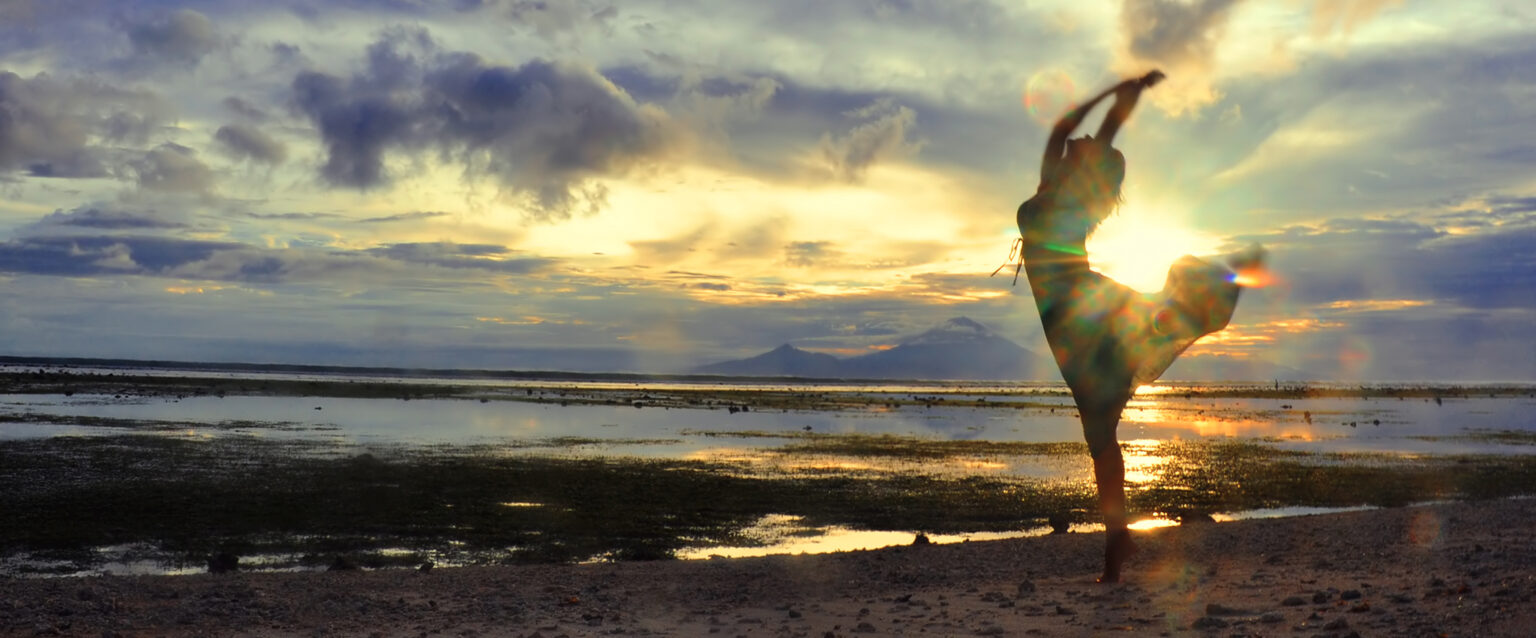 A woman dancing at the beach as the sun goes down over Bali, as seen from Gili, Indonesia.