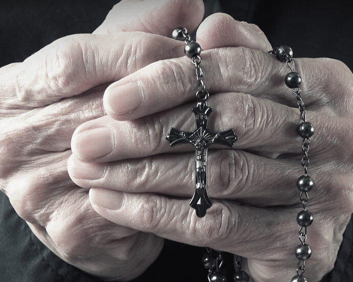 The hands of an elder woman, dressed in black, holding a rosary while praying.