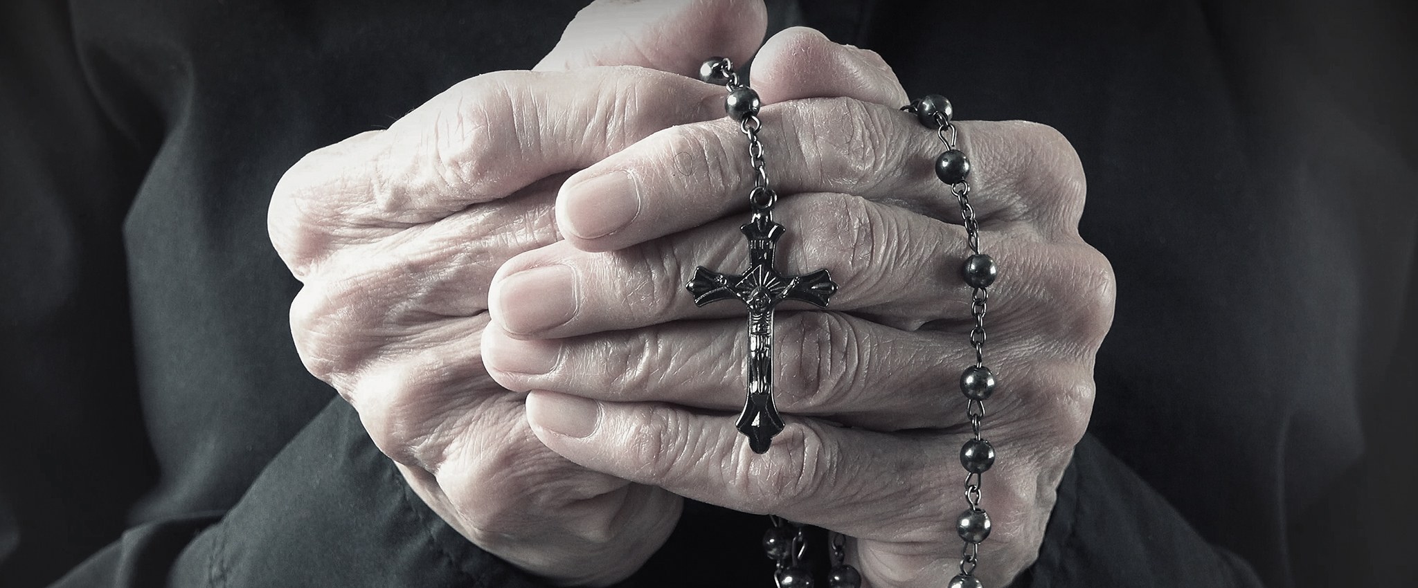 The hands of an elder woman, dressed in black, holding a rosary while praying.