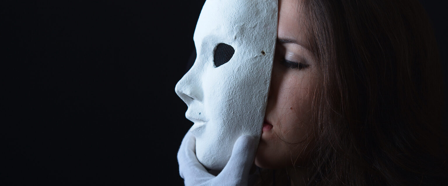 A photo of a brunette woman wearing white cotton gloves and removing a white theatrical mask from her face. The remainder of the image is a black background.