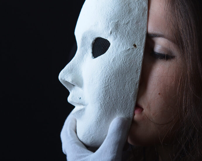 A photo of a brunette woman wearing white cotton gloves and removing a white theatrical mask from her face. The remainder of the image is a black background.
