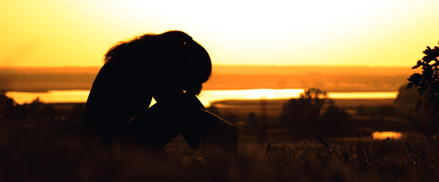 The silhouette of a woman at sunset, sitting in the grass, her head tilted onto her knees.