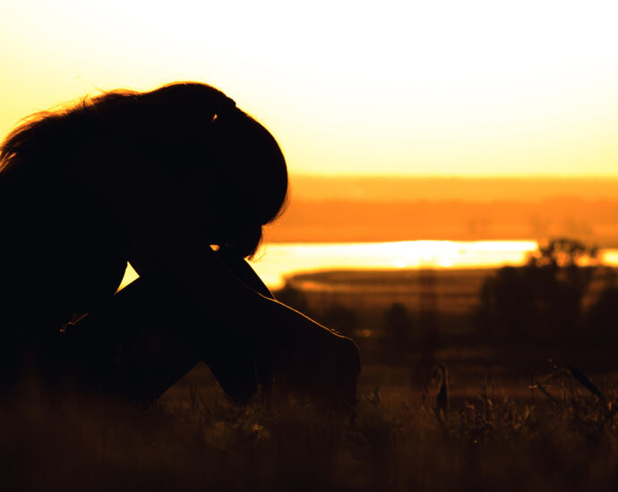 The silhouette of a woman at sunset, sitting in the grass, her head tilted onto her knees.