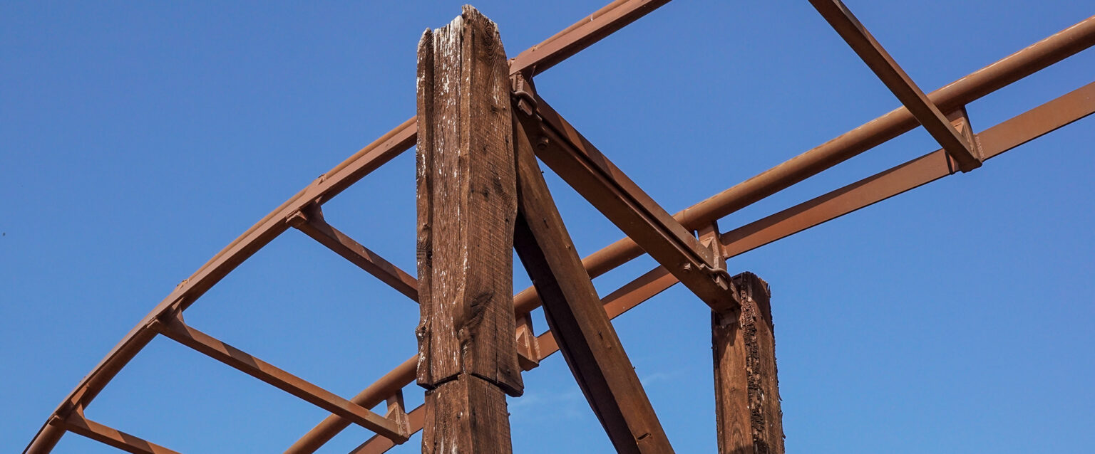 A close-up view of the construction of a large wooden rollercoaster, taken from below. Light-brown steel rails sit atop a wood-encased steel structure.