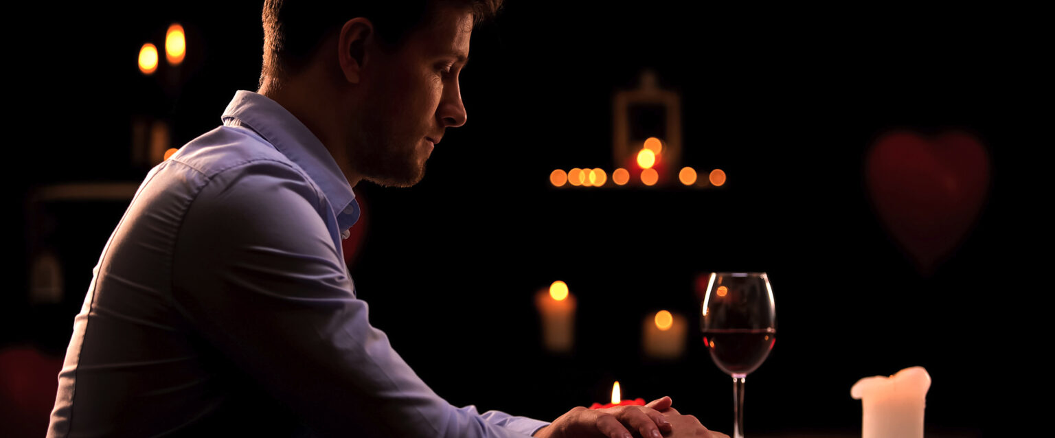 A man sits alone in a darkened restaurant, waiting for his date to arrive. His head is down, and his hands are crossed on the table in front of him. A half-melted candle, a half-empty glass of wine, and his cell phone sit on the table in front of him.