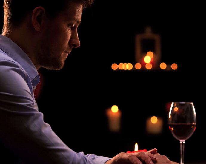 A man sits alone in a darkened restaurant, waiting for his date to arrive. His head is down, and his hands are crossed on the table in front of him. A half-melted candle, a half-empty glass of wine, and his cell phone sit on the table in front of him.