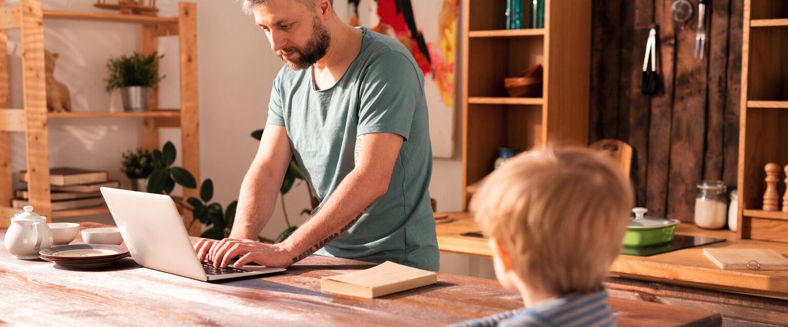 A busy father stands at the kitchen table, working on his laptop, while his lonely son looks at him from the end of the table.