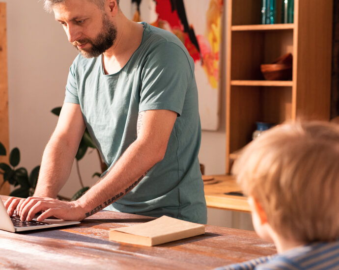A busy father stands at the kitchen table, working on his laptop, while his lonely son looks at him from the end of the table.