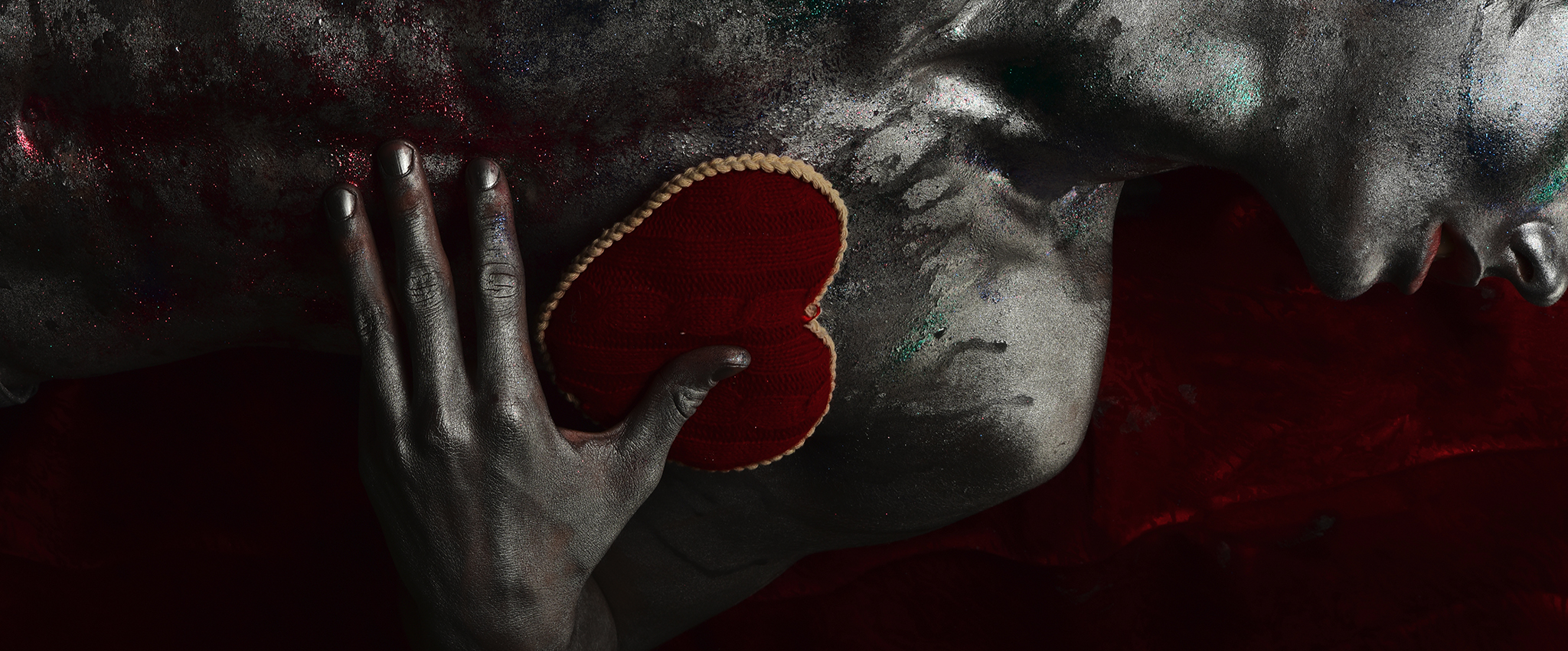 A man, painted silver with splashes of red, lies on a red velvet blanket. His left hand presses a soft, plush red velvet heart, with pearled golden trim, against his chest.