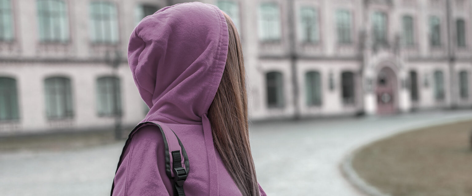 A teen girl with long brown hair, wearing a purple hoodie and matching backpack, looks back over her shoulder at the school she is leaving.