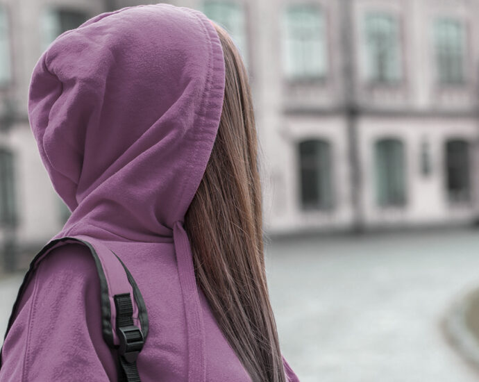 A teen girl with long brown hair, wearing a purple hoodie and matching backpack, looks back over her shoulder at the school she is leaving.