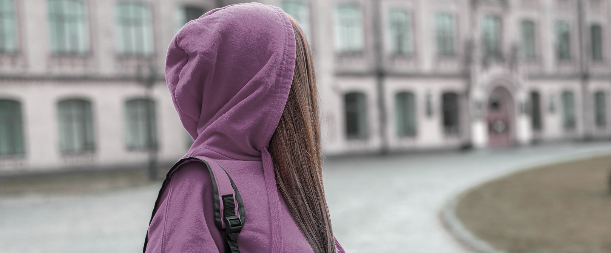 A teen girl with long brown hair, wearing a purple hoodie and matching backpack, looks back over her shoulder at the school she is leaving.