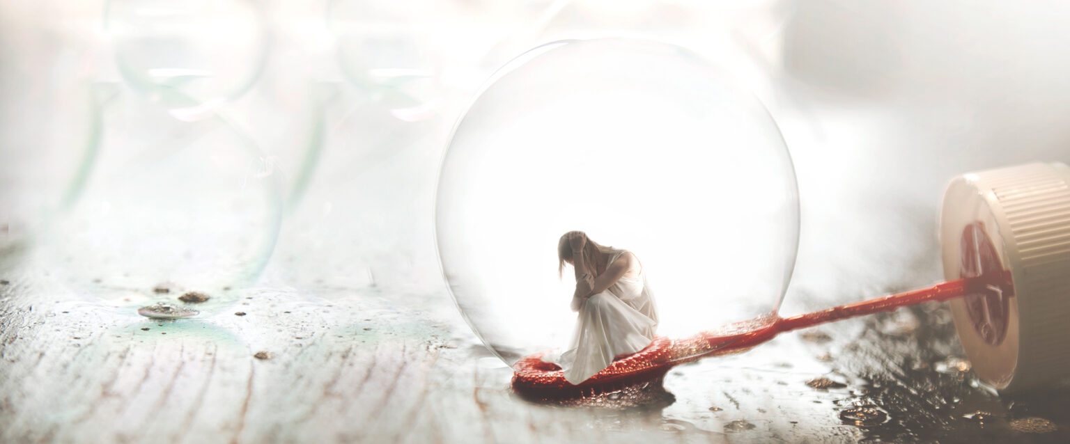 A small woman, in a semi-transparent white dress, sits atop the round end of a red bubble wand, inside a soap bubble.