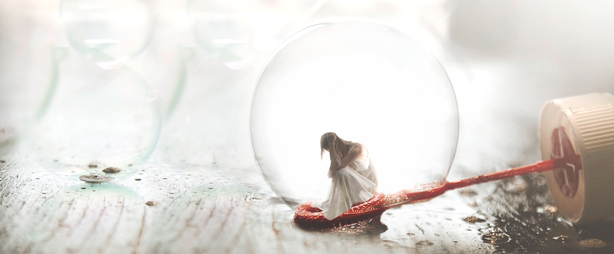 A small woman, in a semi-transparent white dress, sits atop the round end of a red bubble wand, inside a soap bubble.