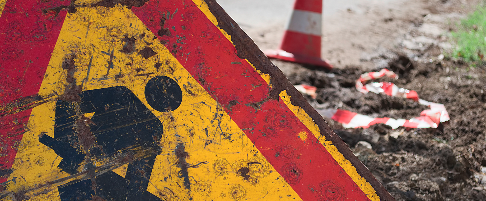 A damaged and rusty yellow and red road sign with a black illustration of a man digging with a shovel sits beside a road, alerting drivers to road work ahead. Beyond the sign, evidence of road work is seen: overturned dirt, a roadside orange caution cone with a wide white horizontal-stripe, and a crumpled section of orange and white caution tape.