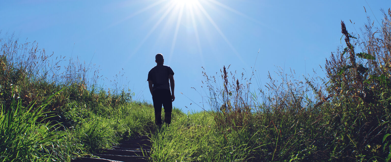 Sun rays silhouette a man as he walks up a grassy hillside stairway toward a blue sky.