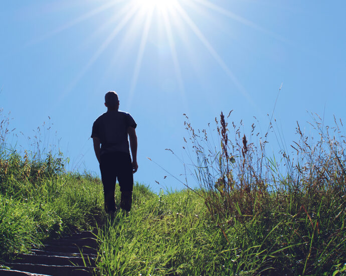 Sun rays silhouette a man as he walks up a grassy hillside stairway toward a blue sky.