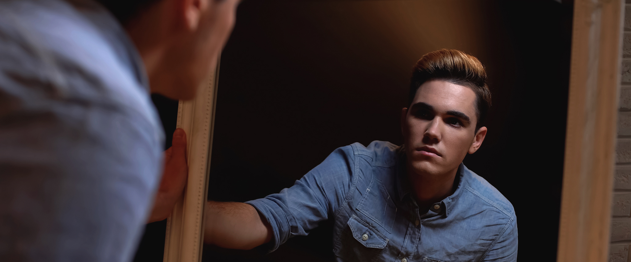A transgender person, experiencing gender dysphoria, looks into a tall, wood-framed mirror.