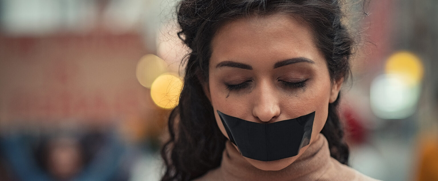 A woman with black hair stands, eyes closed, with a large piece of black tape over her mouth. In the blurred background, protestors holding signs can be seen.