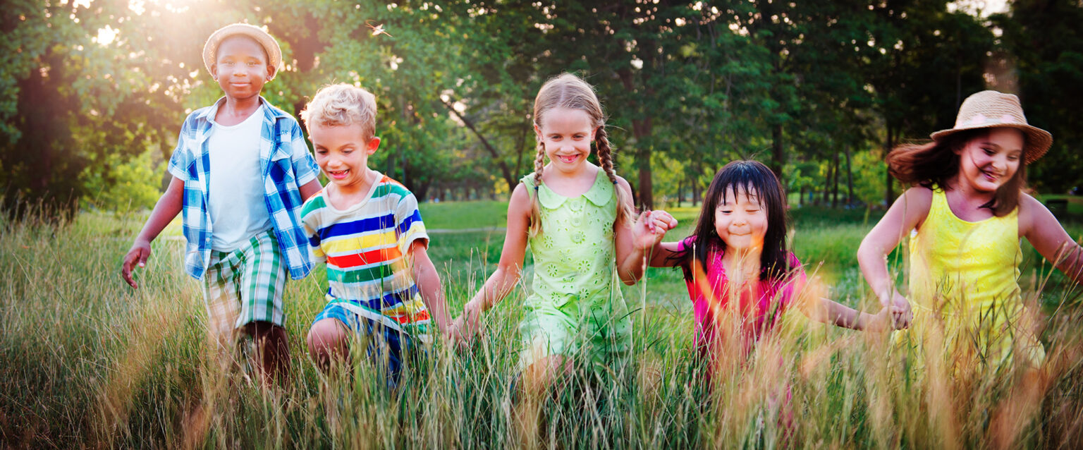 A diverse group of five children hold hands as they run across a field, lined wth trees, into the tall grass, in the foreground.