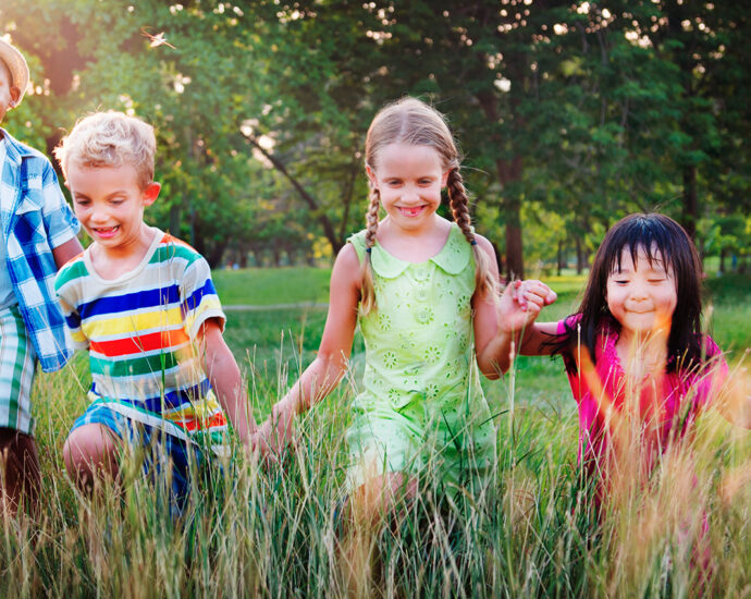 A diverse group of five children hold hands as they run across a field, lined wth trees, into the tall grass, in the foreground.