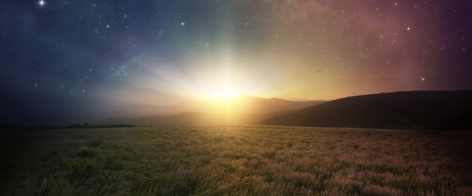 A beautiful sunrise peeks out above distant mountains and shines across the grass-covered plain. The sky above it is filled with the lush stars of the night sky. Its color, visible through the sun's rays, fades from dark blue on the left to mauve on the right.