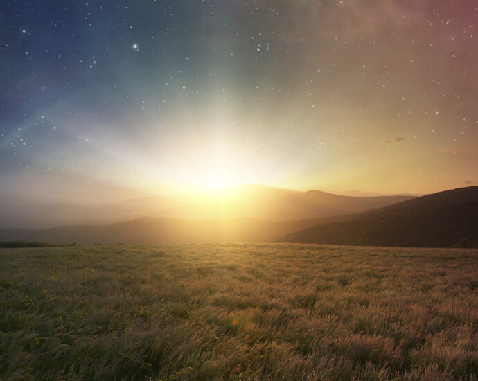 A beautiful sunrise peeks out above distant mountains and shines across the grass-covered plain. The sky above it is filled with the lush stars of the night sky. Its color, visible through the sun's rays, fades from dark blue on the left to mauve on the right.