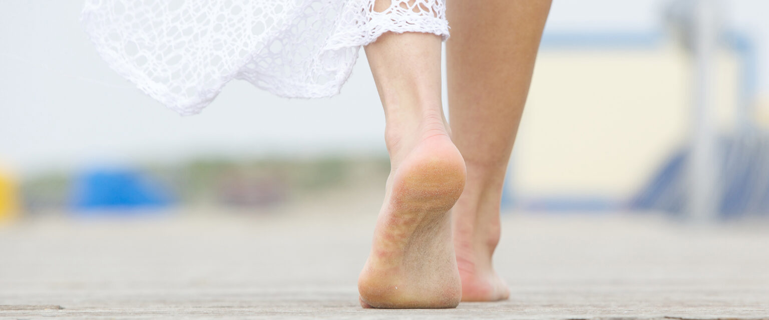 A close-up, low-angle photograph of a barefoot woman, in a white lace dress, walking away.