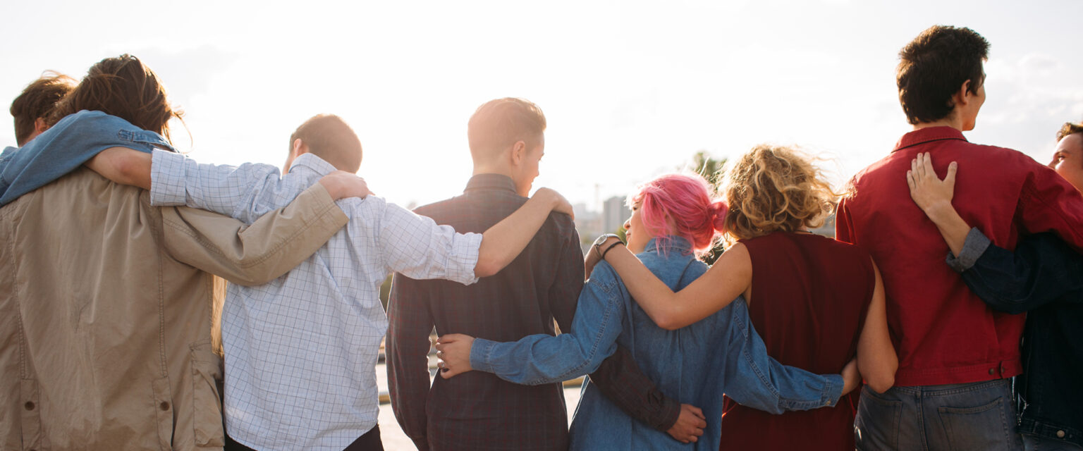 Back view of a diverse group of 8 young people, with their arms around the people on each side of them. Unity and support. Friendship together.